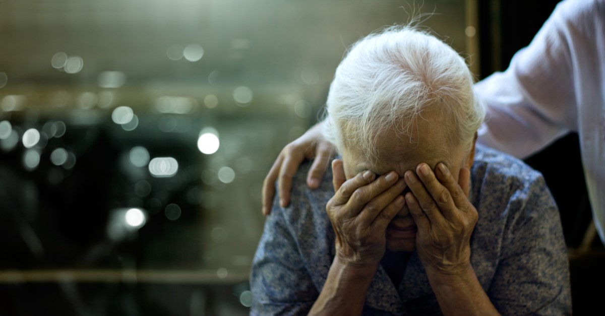 Eine ältere Frau hat ein gesundheitliches Problem, ihr Kopf wird von ihren Armen gestützt und eine Person hält ihre Schultern.