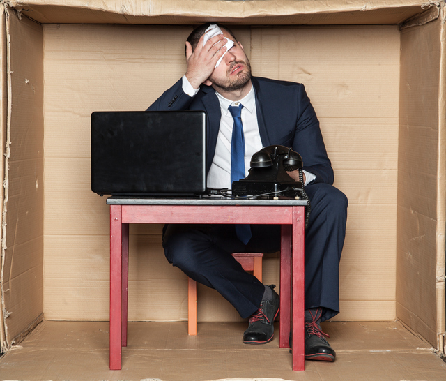 Geschäftsmann an einem Schreibtisch in einer Box sitzend, beengte Räumlichkeiten, Arbeitsstress und erhöhte Arbeitsbelastung, Computer und Telefon, Mann wischt sich die Stirn