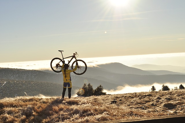 Eine Radfahrerin hebt ihr Fahrrad über den Kopf, im Hintergrund sind Berge im Nebel zu sehen.