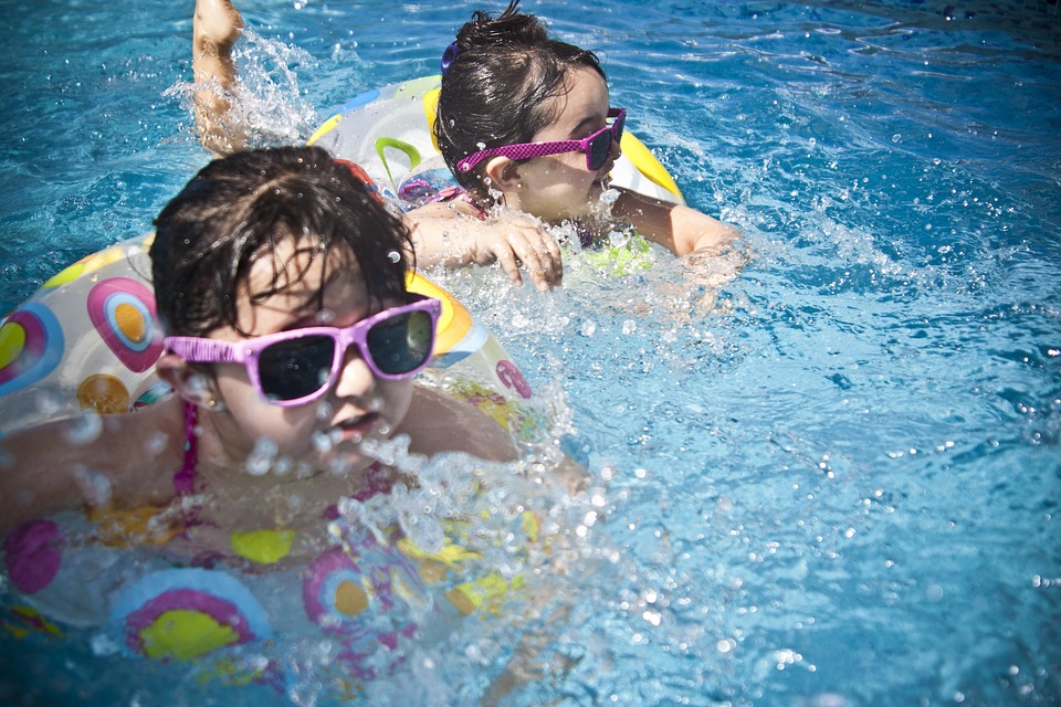 Zwei Kinder schwimmen im Wasser. Sie tragen Sonnenbrillen mit rosa Gestellen und schwimmende Fahrräder.