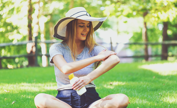 Eine junge Frau trägt Sonnencreme auf, während sie auf einer Wiese sitzt und einen Hut trägt, um ihren Kopf zu schützen