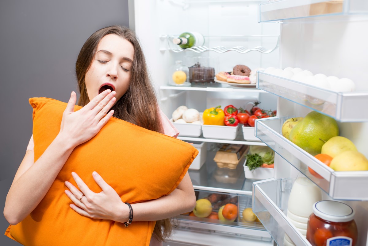 Eine Frau, die neben einem Kühlschrank schläft, veranschaulicht, dass die Essgewohnheiten einen Einfluss auf die Schlafqualität haben.