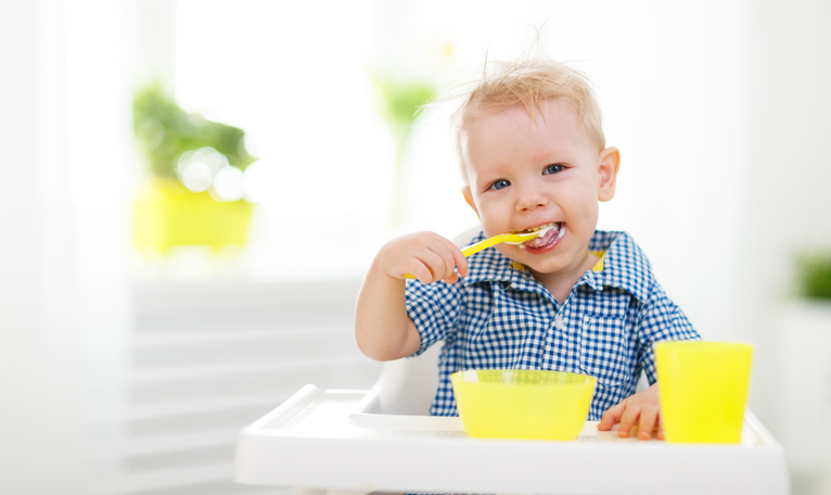 Ein kleines Kind, das in einer Babyschale sitzt und sich selbst füttert. Gelber Löffel, Tasse und Schüssel