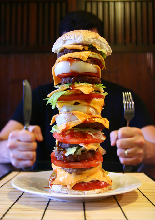 Der Mann sitzt am Tisch, auf einem Teller hat er einen großen Hamburger, eine Megaportion