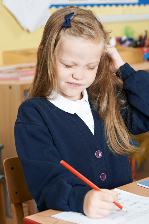 Ein kleines Mädchen sitzt in der Schule mit einem juckenden Kopf, wahrscheinlich Läuse im Haar, blondes Haar, sitzt und schreibt