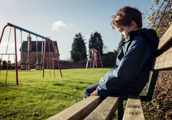 kleiner Junge sitzt alleine auf einer Bank, ist traurig, erlebt negative Emotionen, Spielplatz, Gras, Schaukel
